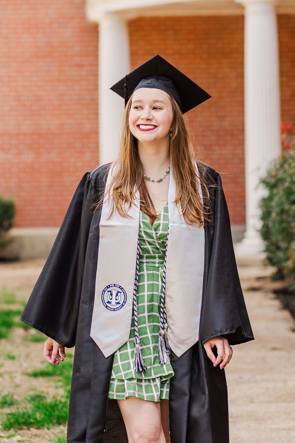 Graduate wearing cap and gown with green dress underneath standing by brick building.