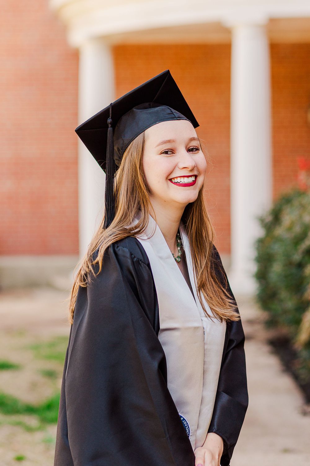 Graduate in black cap and gown with gray stole posing by white columns.