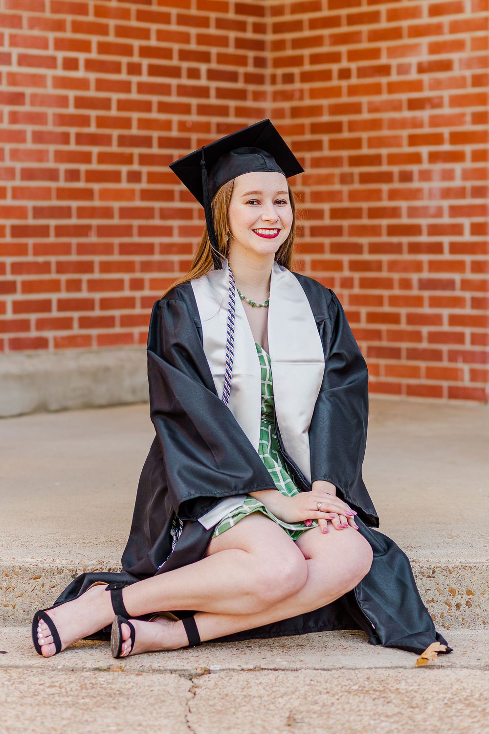 Graduate in cap and gown sitting cross-legged on concrete with brick wall backdrop.