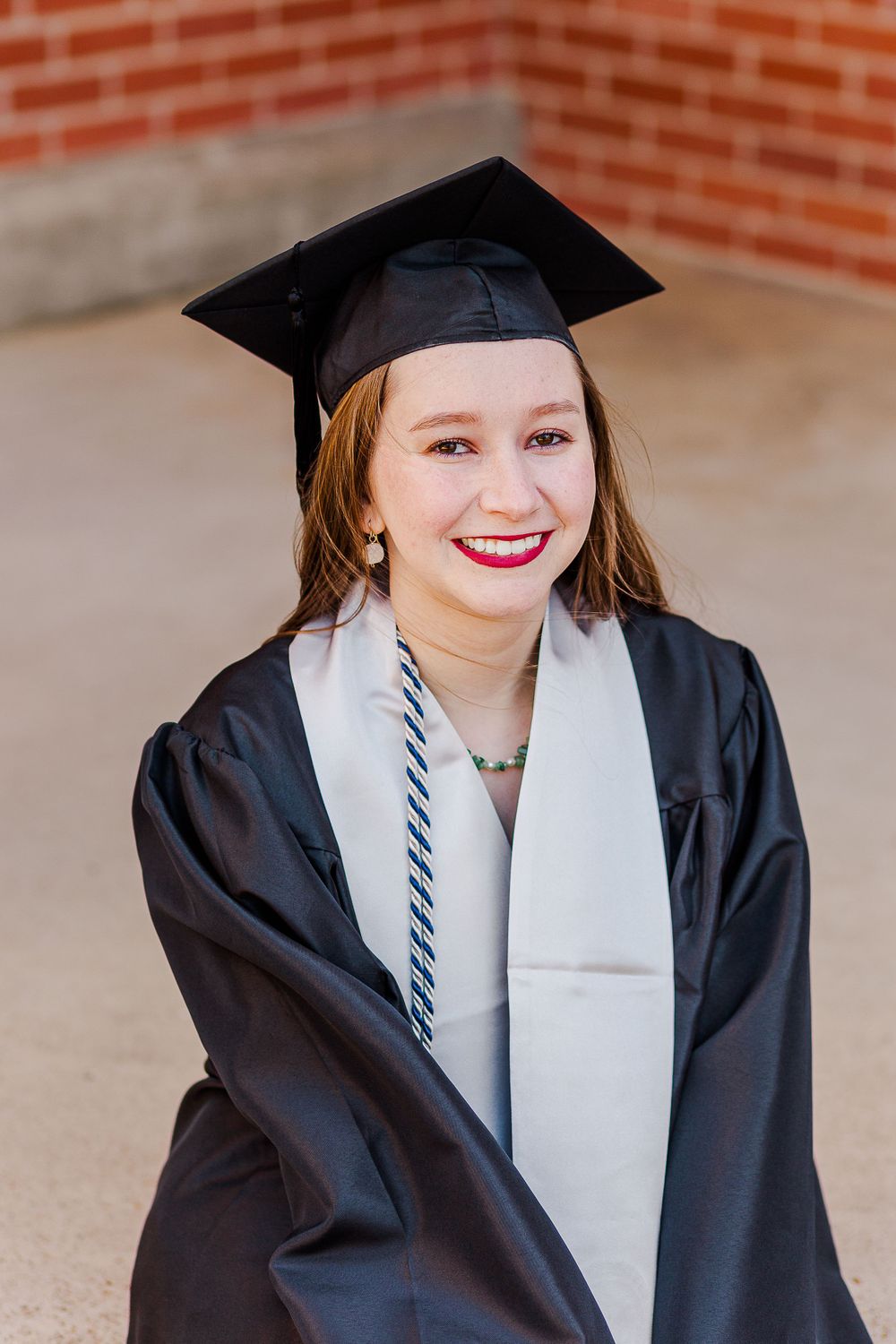 Graduate in traditional cap and gown smiling against brick background.