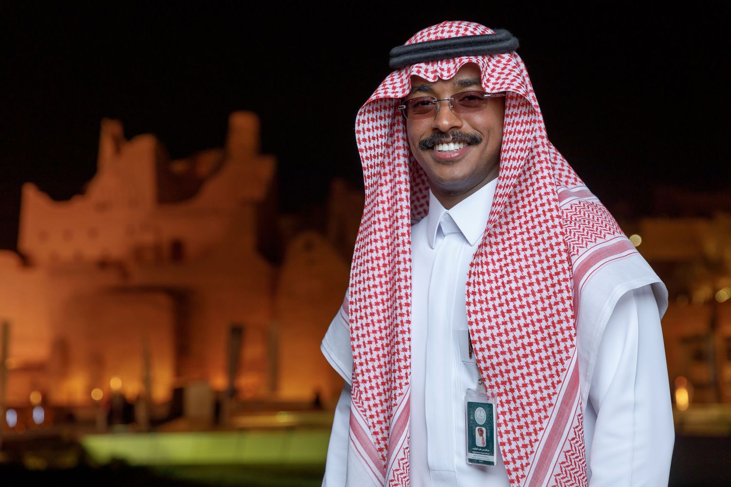 Person in traditional white thobe and red checkered headscarf smiling at night with historic building behind.