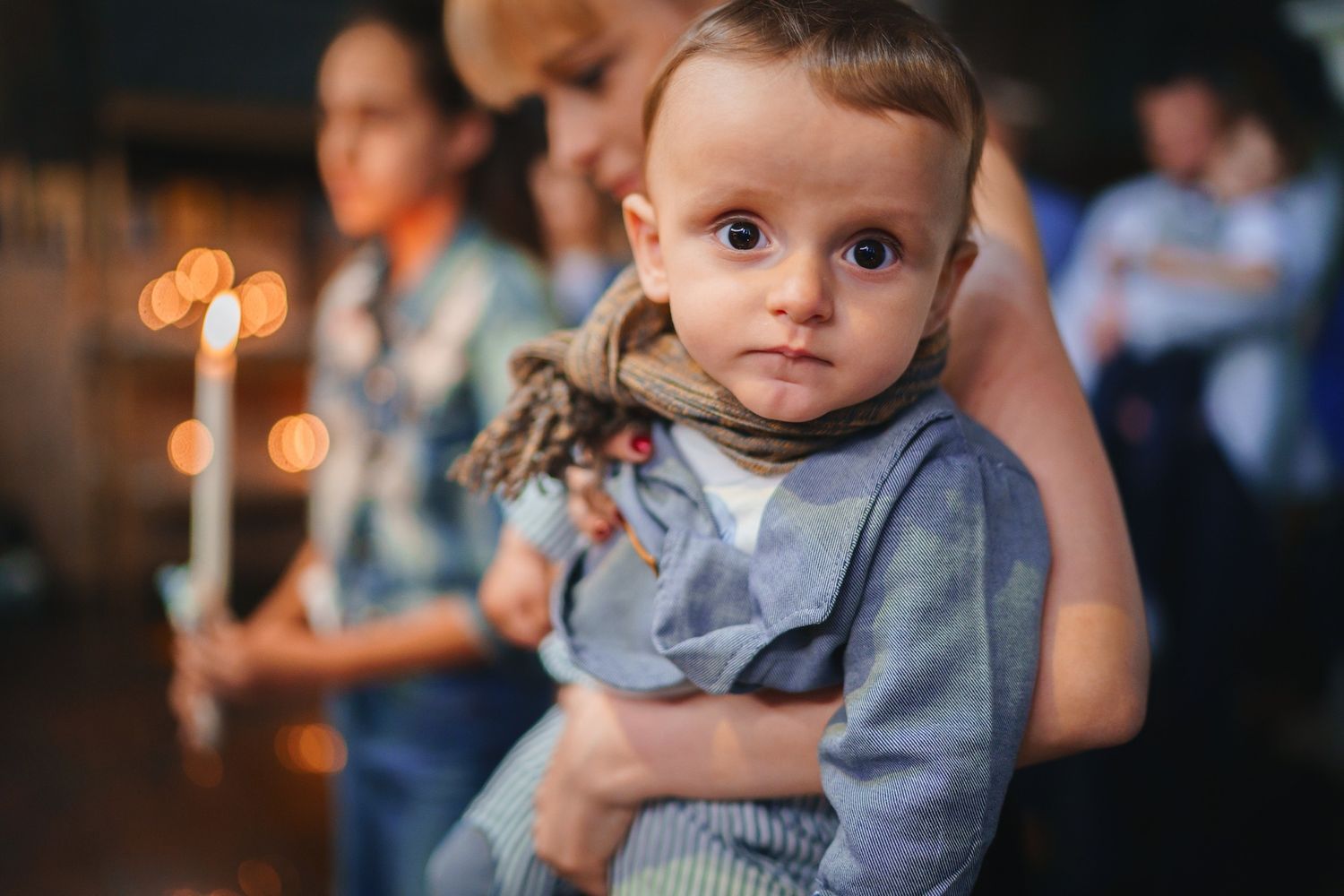 Baby in gray outfit looks towards camera during candlelit ceremony.