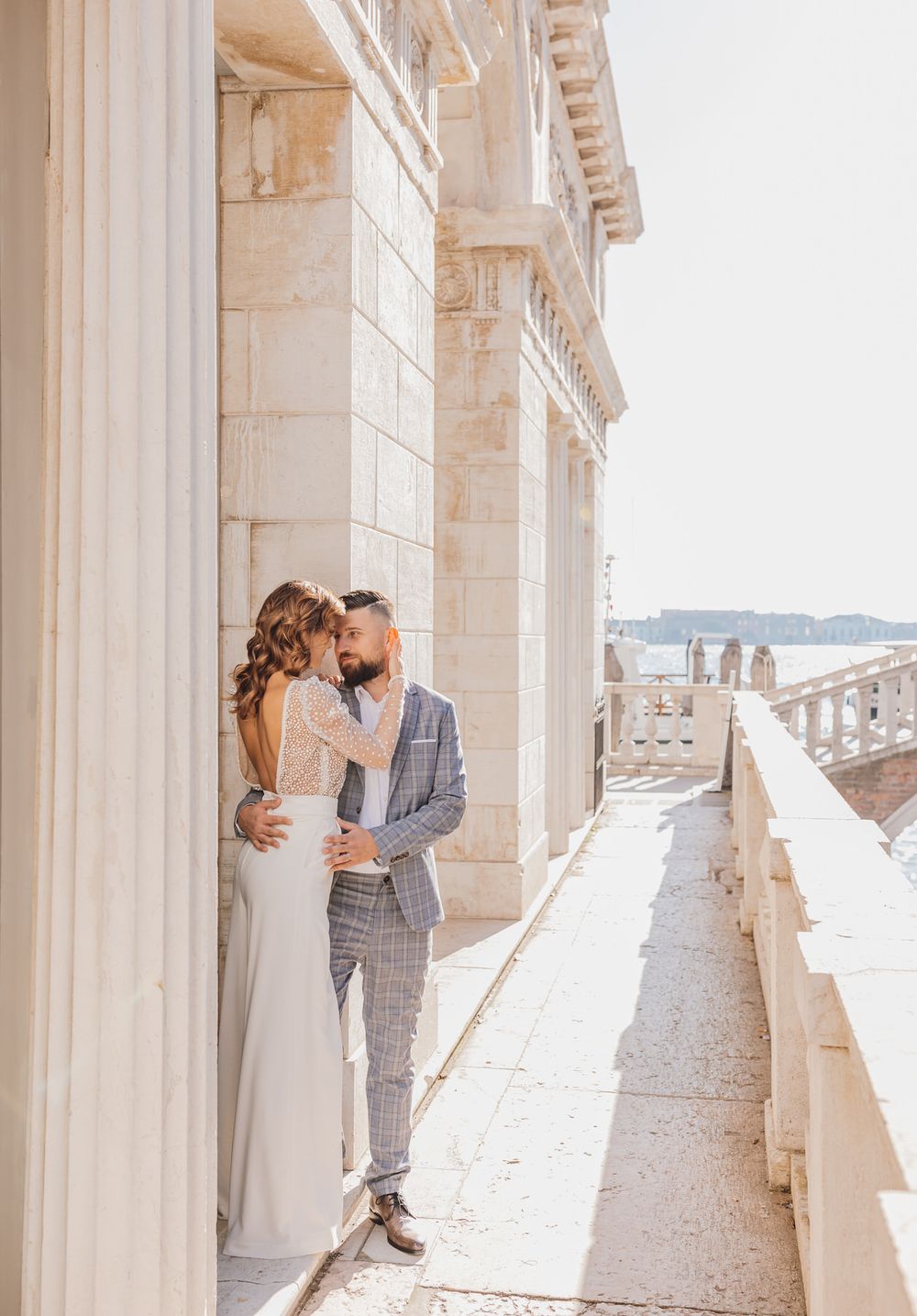 Romantisches Hochzeitspaar umarmt sich auf weißem Steinbalkon mit malerischem Meerblick. Venedig