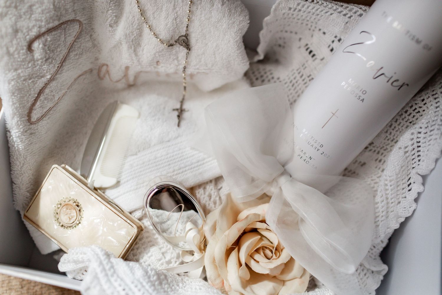 Wedding details flatlay with lace, rings, and dried rose on white textile.