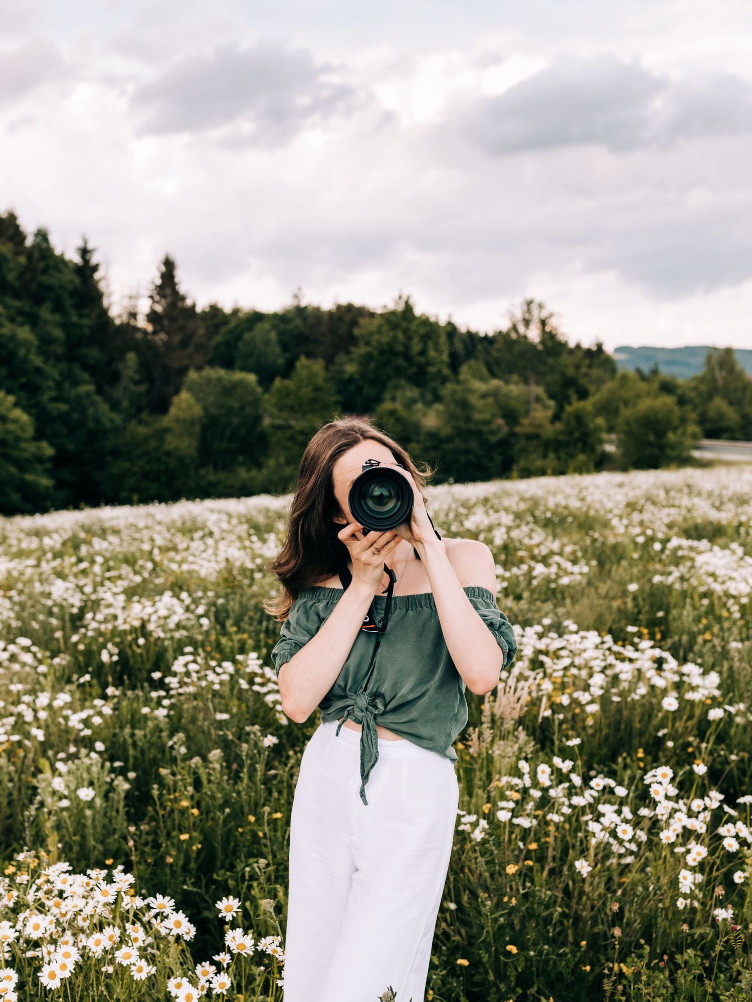 A photographer in an off-shoulder top captures images in a meadow filled with white wildflowers.