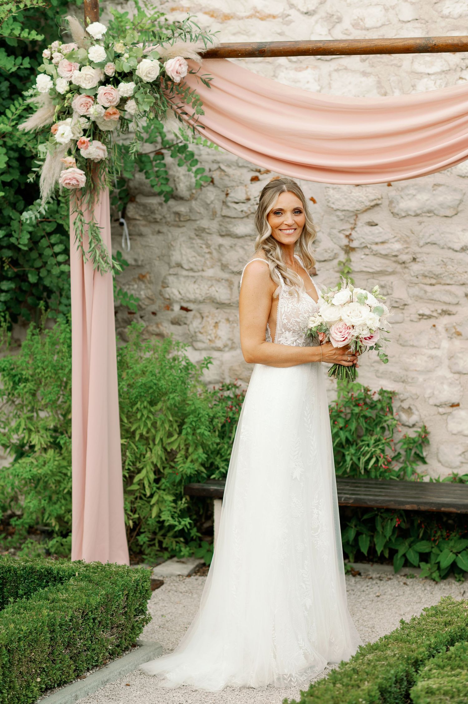 Bride in lace wedding dress poses with bouquet under floral arch with blush draping.