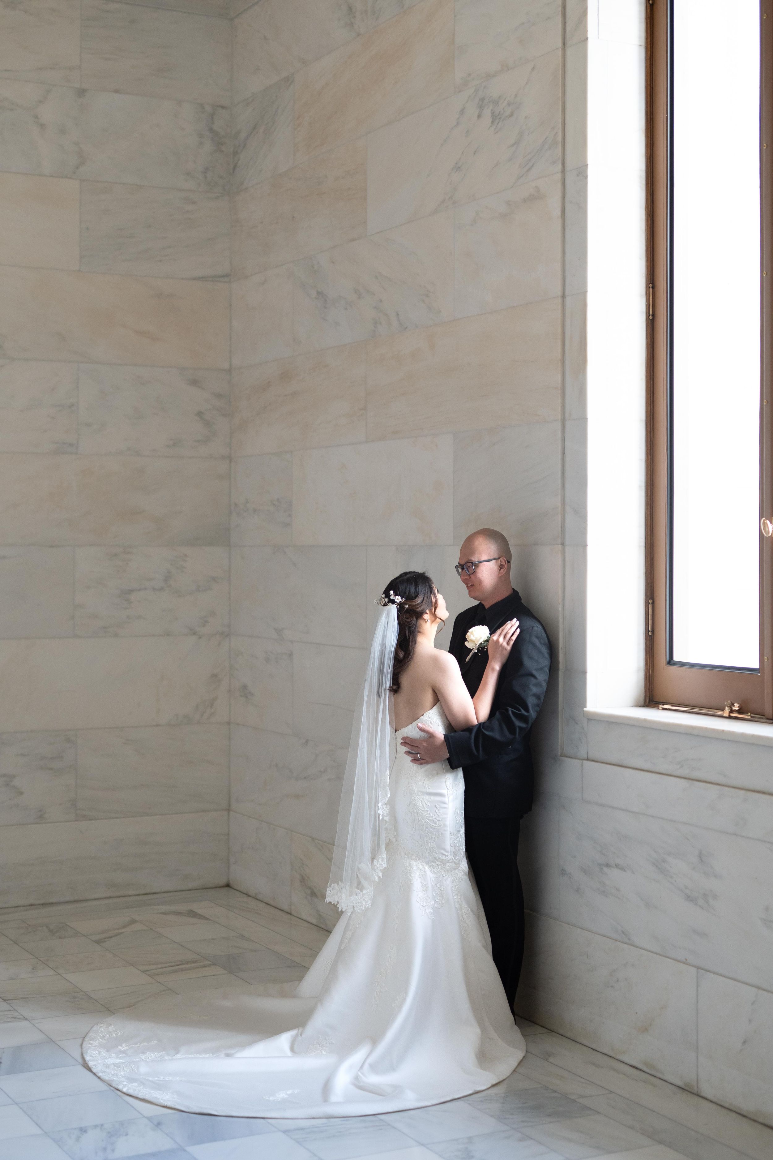 Bride and groom share intimate moment against marble walls near window.