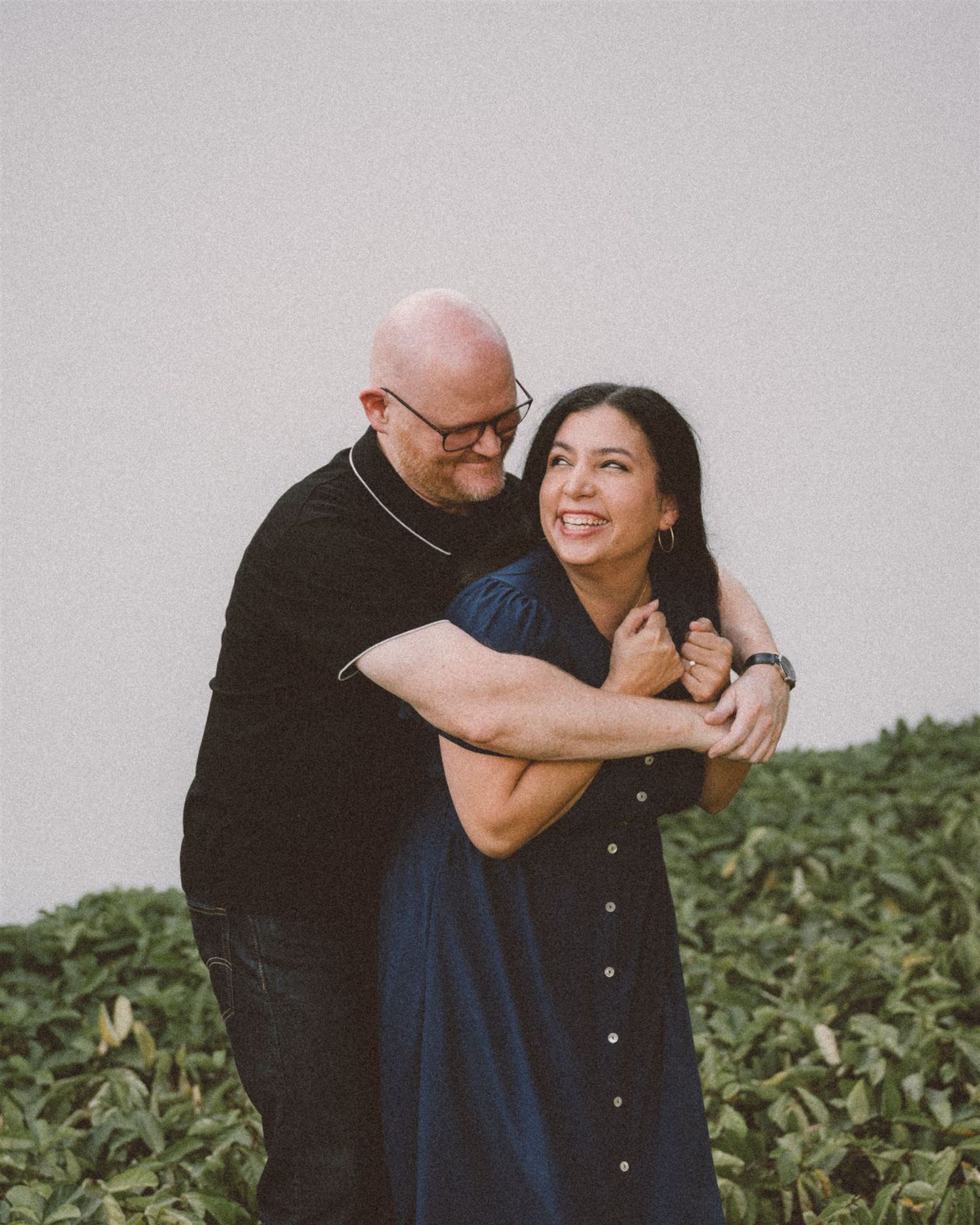 A romantic couple shares a warm embrace against a light colored wall with greenery in the background.
