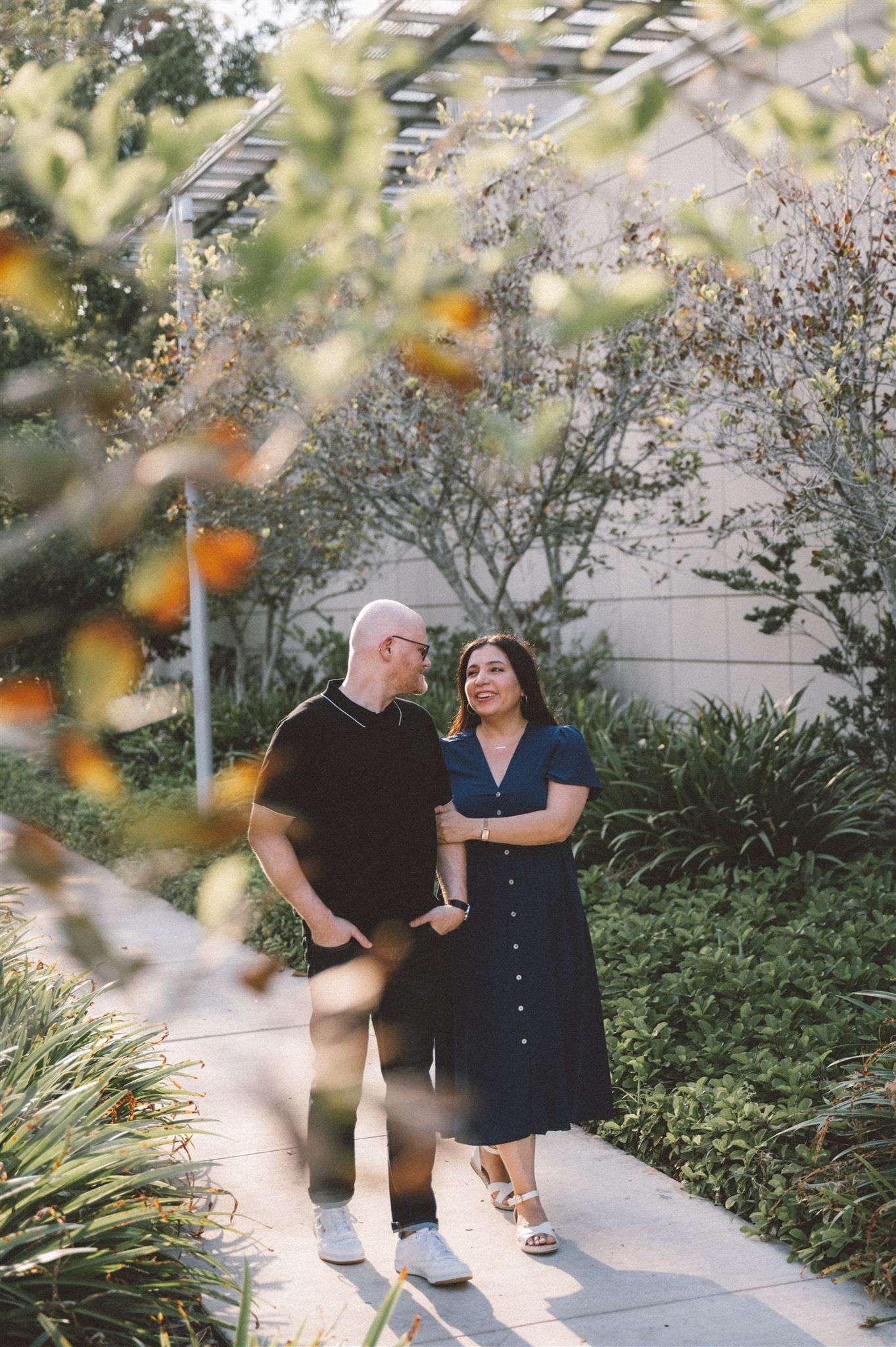 A couple poses together on a pathway surrounded by blooming plants and trees.