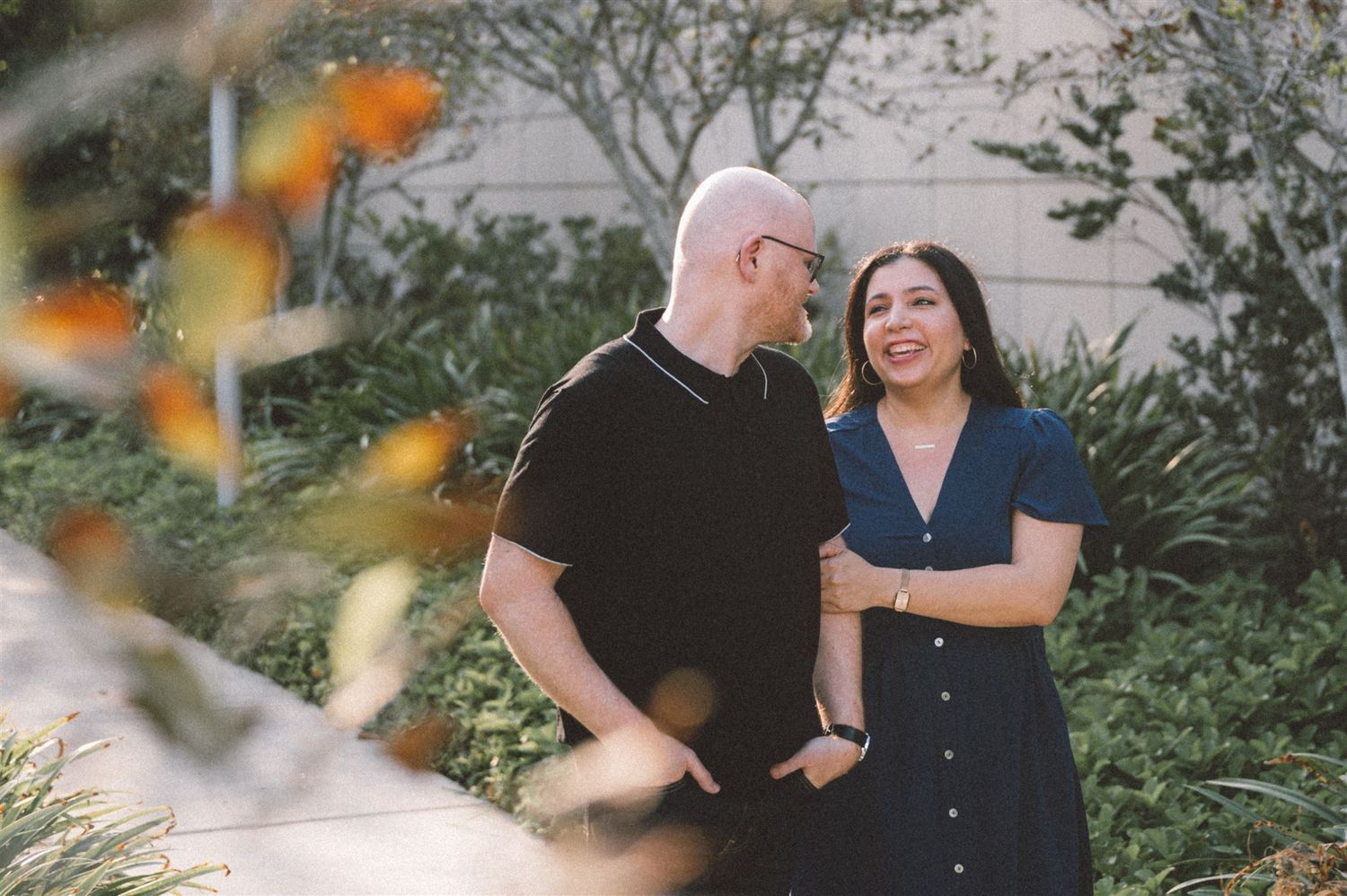 A couple shares a laugh while standing on a garden path framed by autumn leaves.