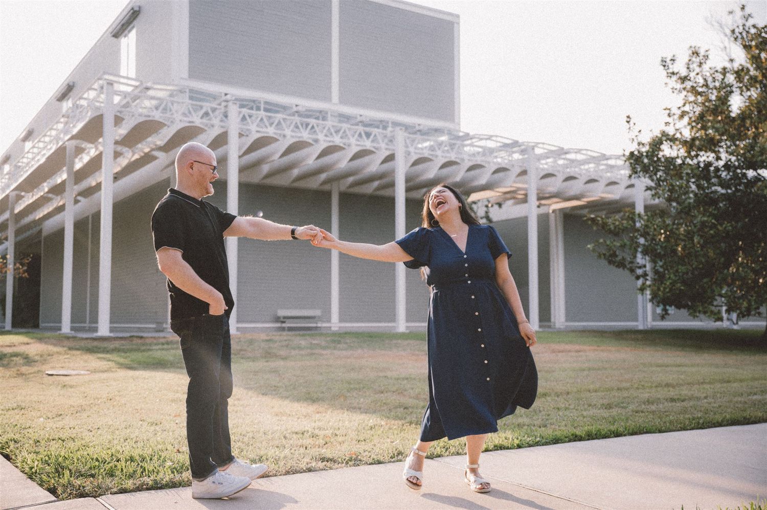 A couple dances playfully in front of a modern building with a white pergola.