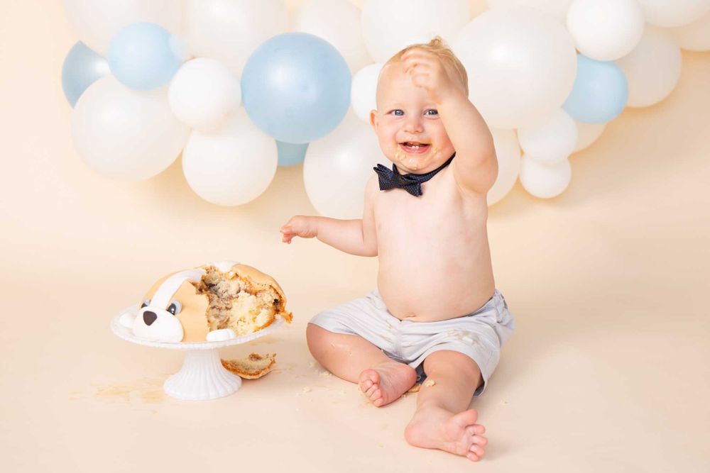 Laughing baby in diaper with bow tie poses next to smashed cake.