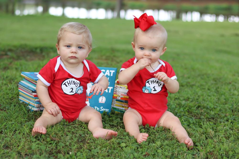 Two babies in matching red outfits sitting on grass with books.