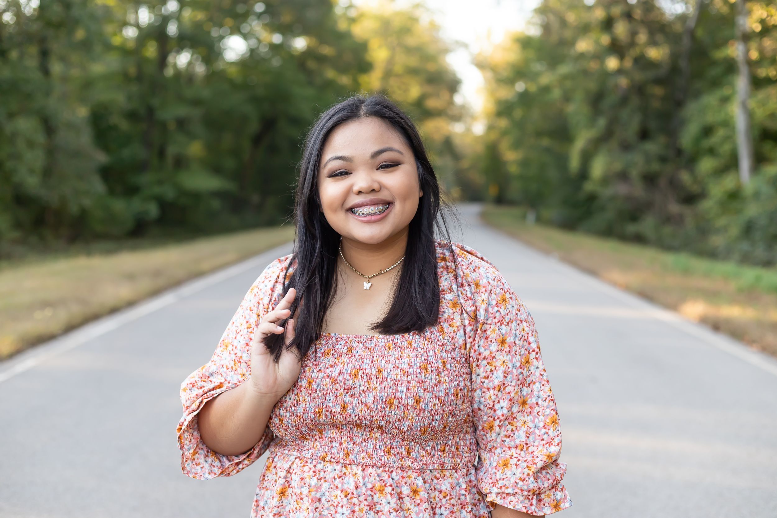 Person wearing a pink floral dress poses on a tree-lined road during golden hour.