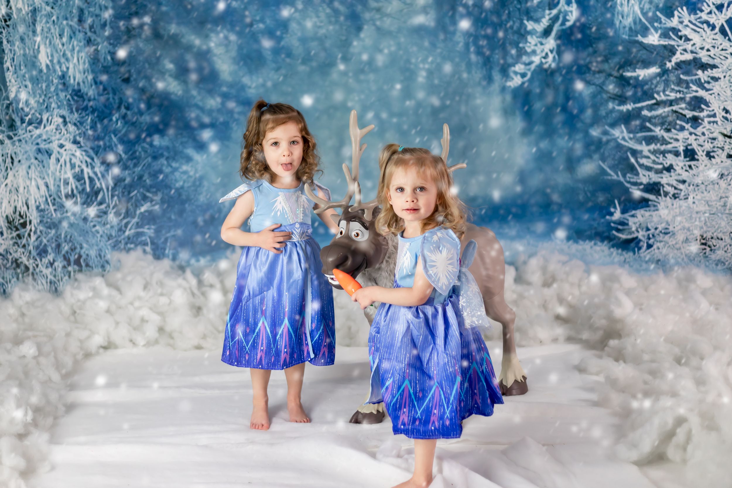 Two children in matching blue dresses dance in winter wonderland setting with snowy backdrop.