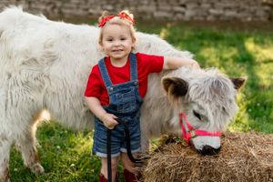 White pony and small child in overalls posing together on hay bale.