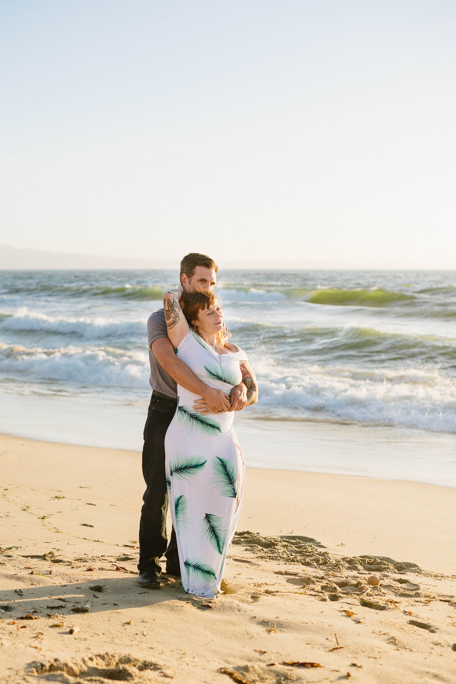 A couple shares an intimate moment on a sandy beach with rolling waves in the background.