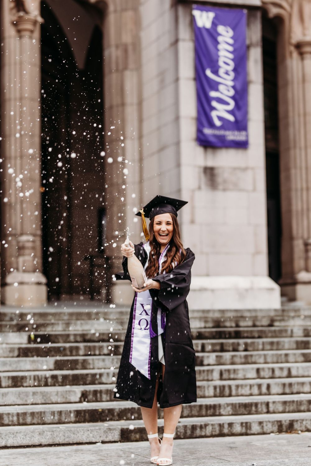 Graduate celebrating on university steps with purple welcome banner and falling confetti in background.
