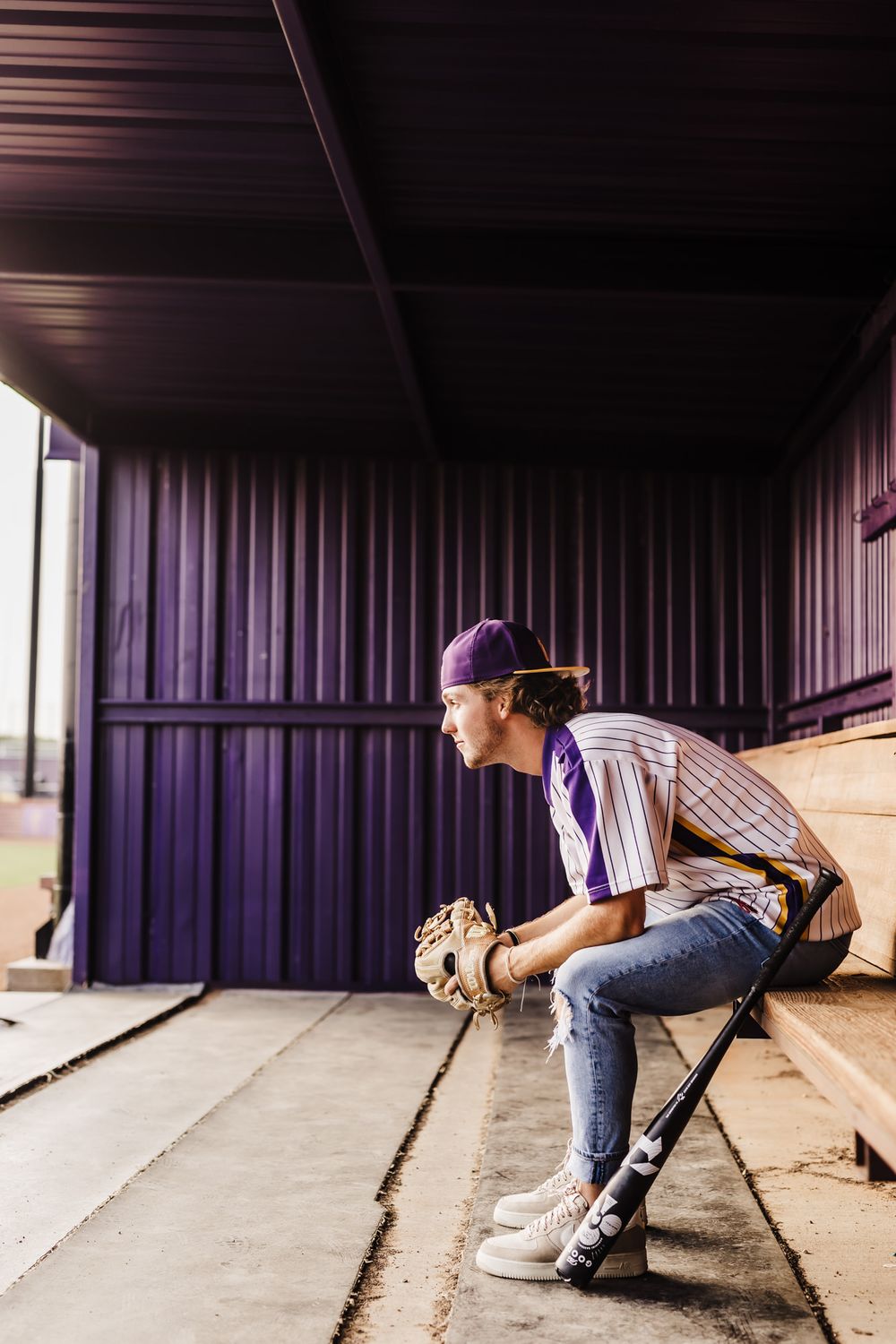 A baseball player sits in dugout wearing purple uniform with baseball glove.