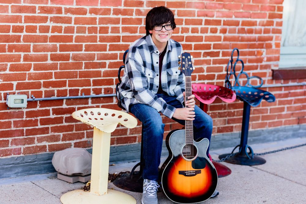 Musician with acoustic guitar sitting on stool against brick wall background.