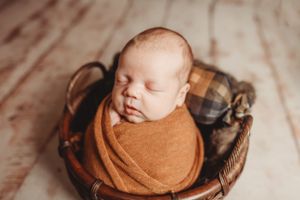 Newborn baby wrapped in brown blanket resting peacefully in wooden basket.