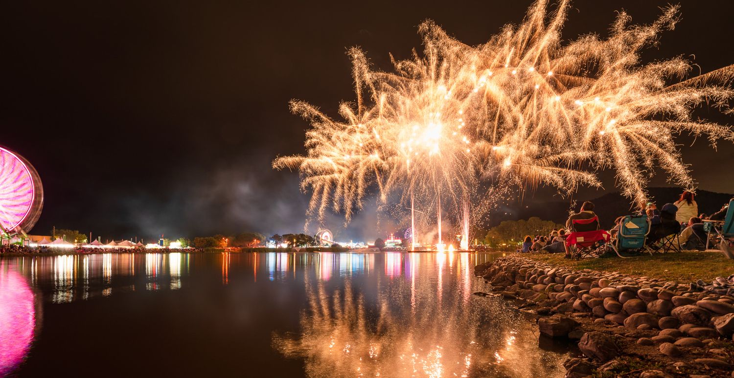 Colorful fireworks display reflects on calm lake waters at night with illuminated shoreline and buildings visible.