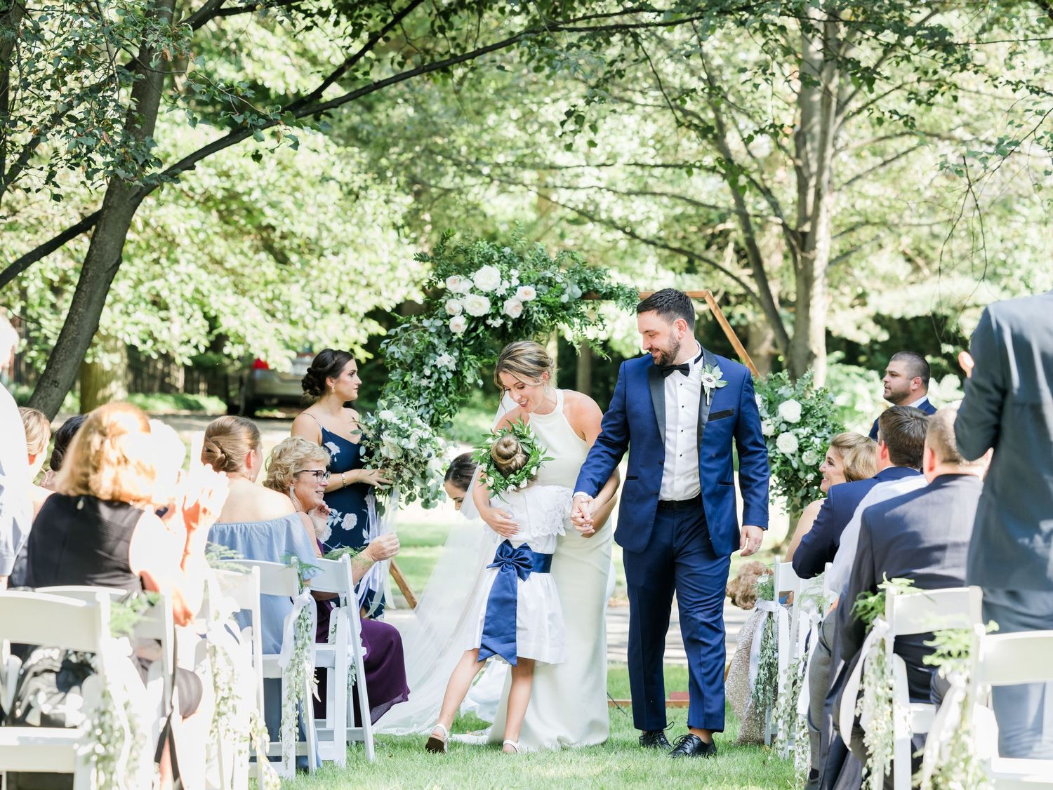 bride and groom walking down isle in yard and get hug from flower girl