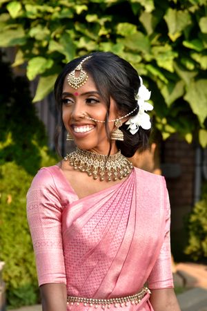 Traditional South Asian bride in pink saree with ornate jewelry and flowers against green foliage.