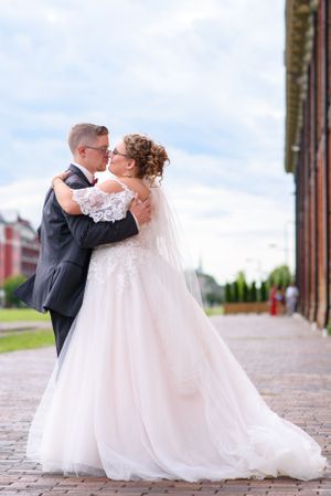 Newlyweds embrace on cobblestone street with historic building in background.