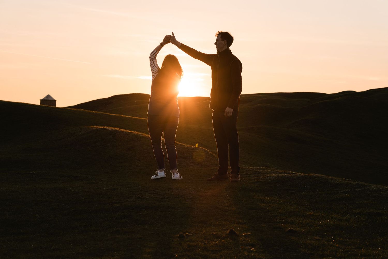 A young couple dancing during golden hour, across open fields. A silhouette of them, with the sun shining through the gap between them.