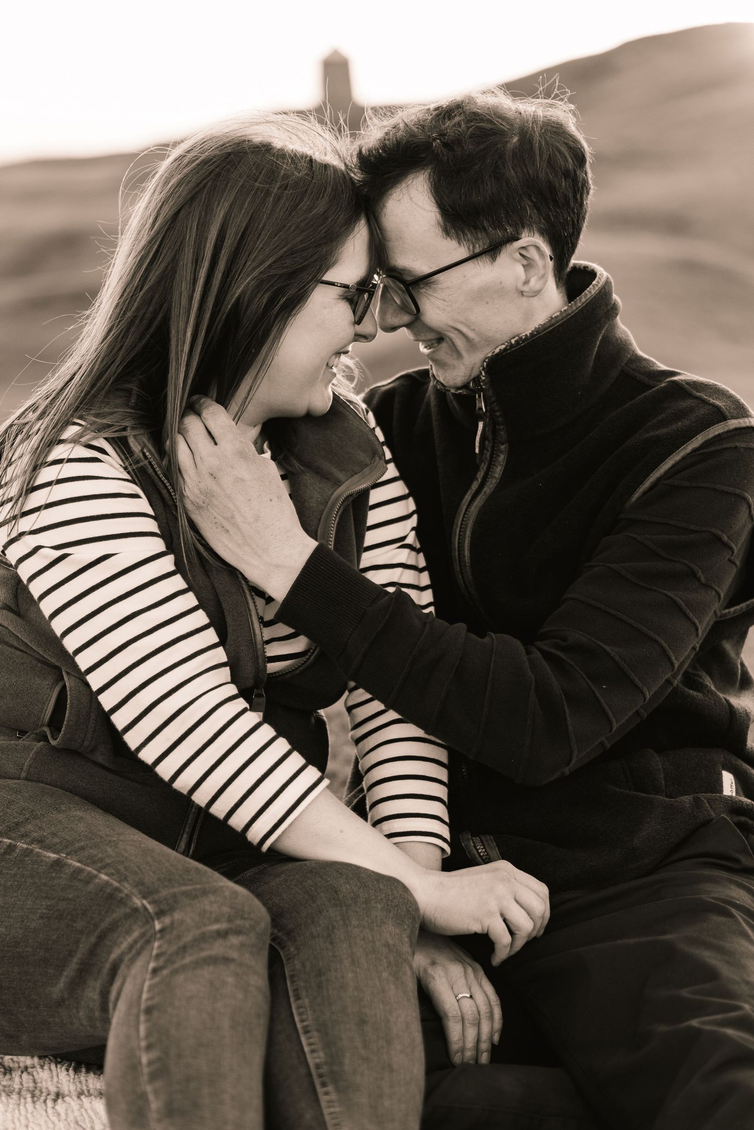 A black and white image of a young couple sat down. They have their heads together and he is touching her neck.
