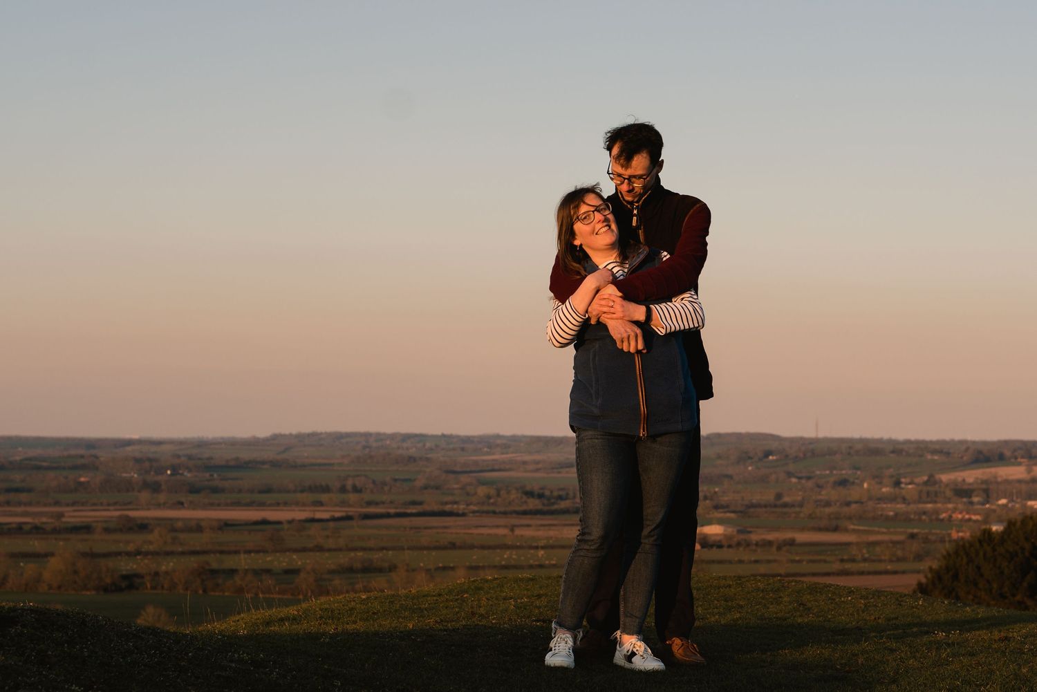 A young couple having an embrace. Him behind her with his arms wrapped around her. Golden hour and open countryside behind them.