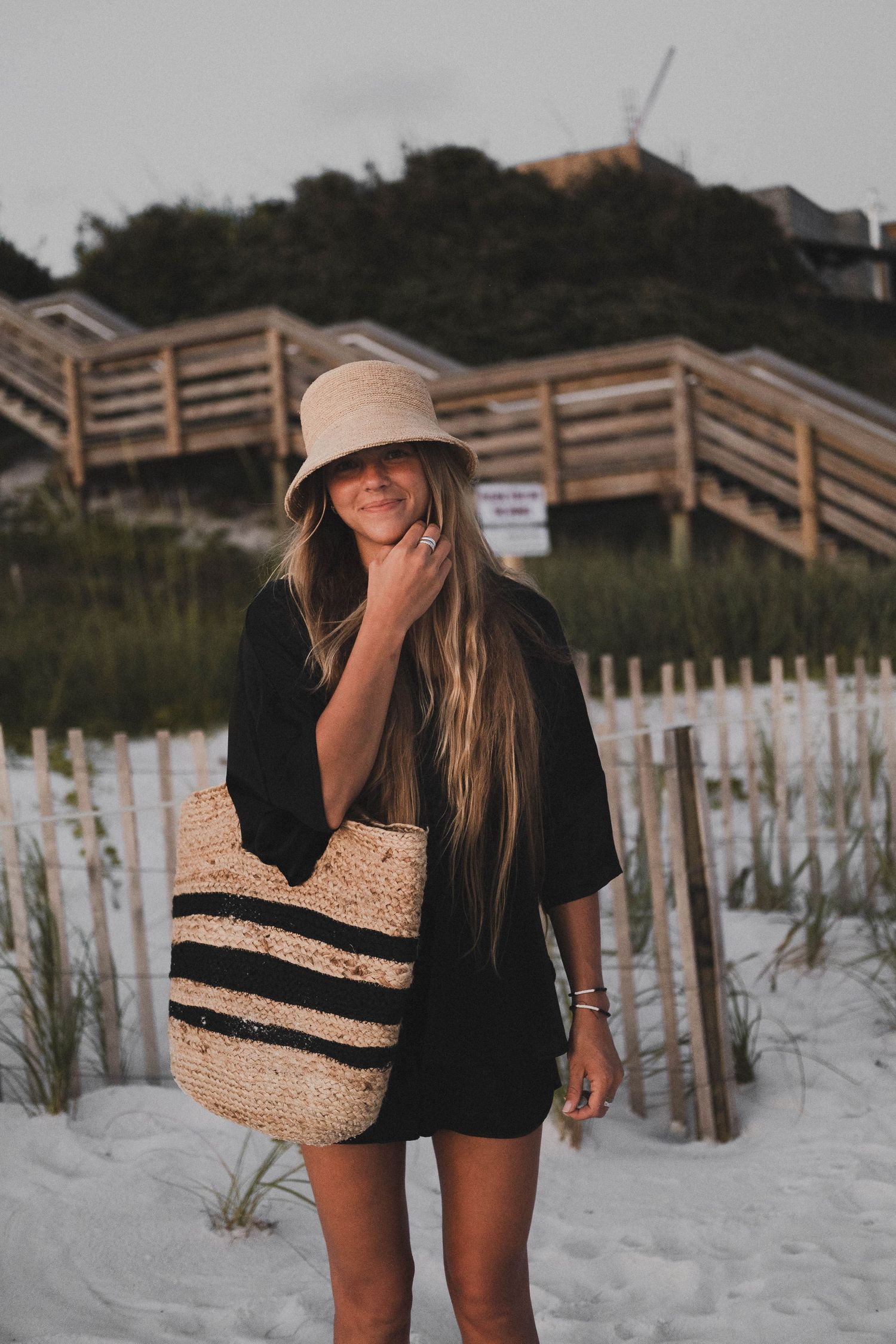 Person walking on white sandy beach with straw bag and sun hat at dusk.