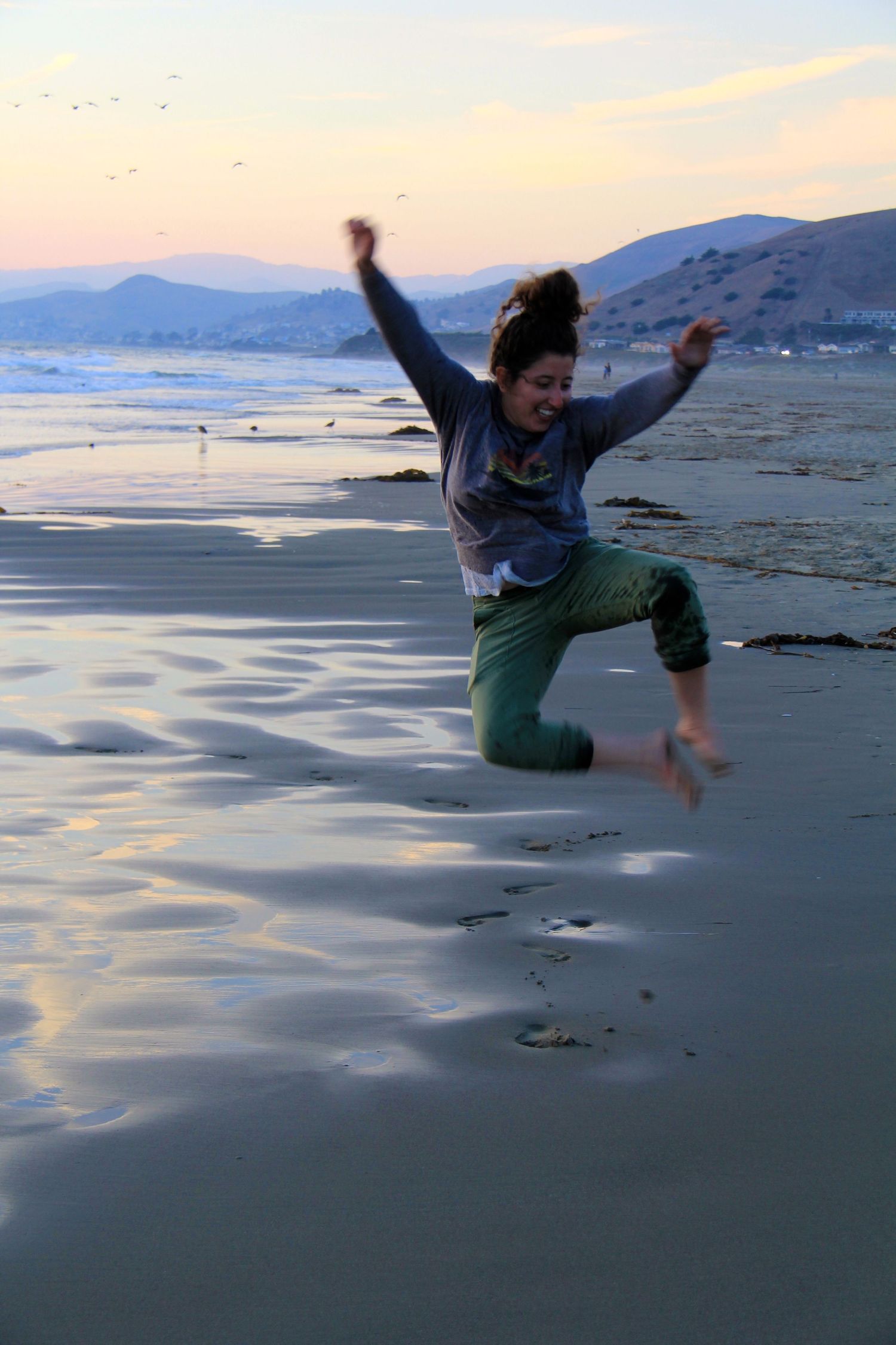 Figure leaping joyfully at sunset on a reflective beach with mountains in background.