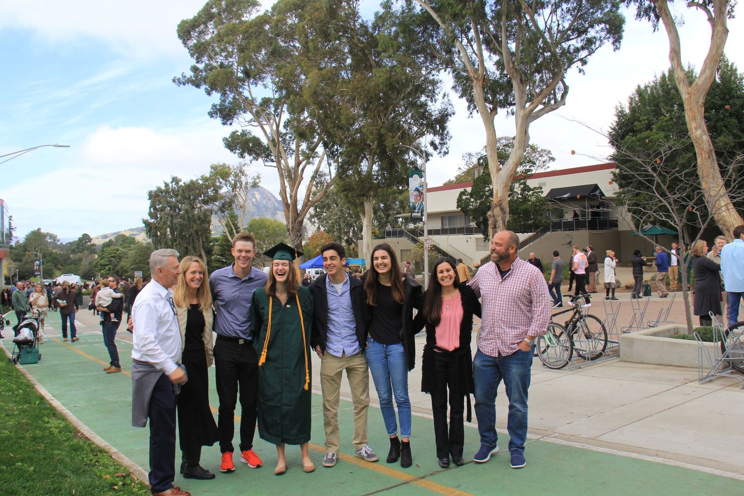 Group photo of graduates and family celebrating on a tree-lined campus pathway.