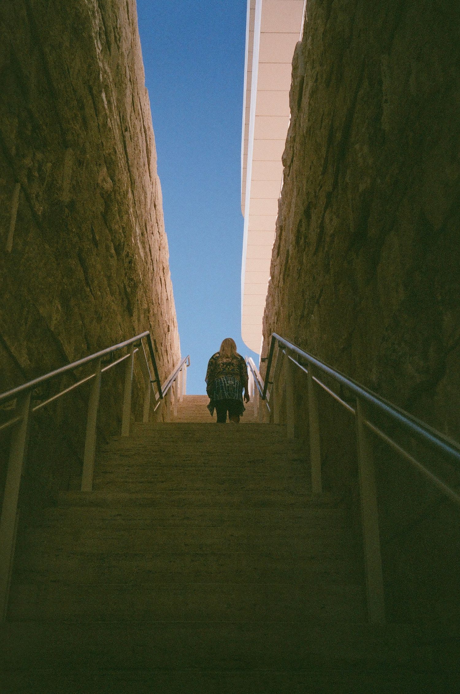 A silhouetted figure ascending concrete stairs between high walls under blue sky.