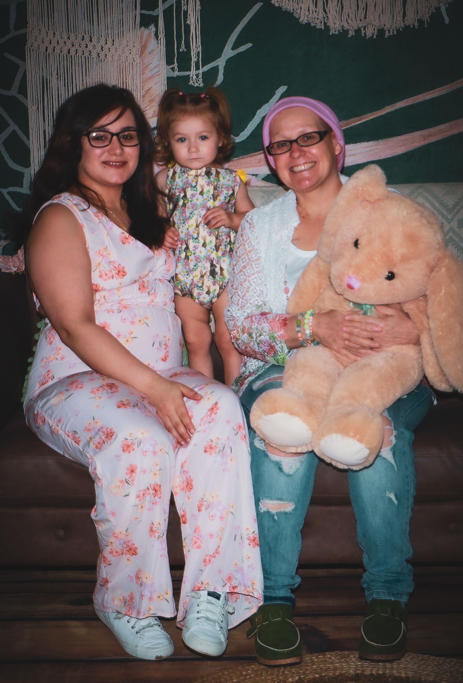 Family portrait with stuffed bear on couch against macrame backdrop with tropical elements.
