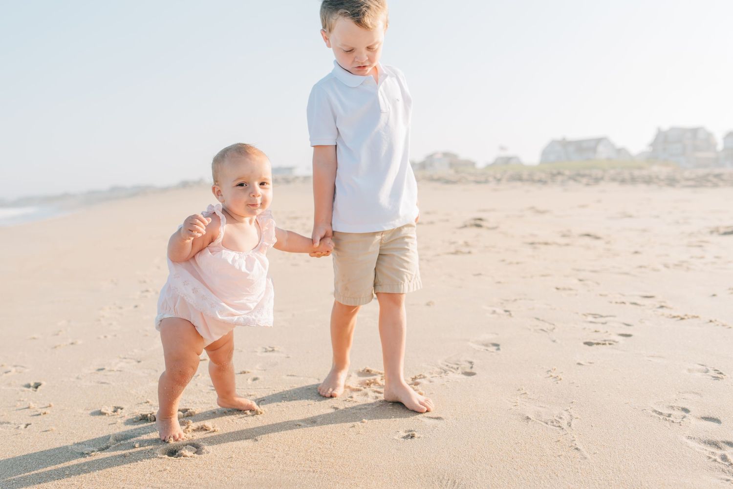 Big brother holds sisters hand while walking down the beach on a July night.