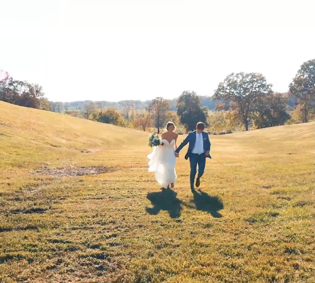 Newlywed couple runs hand-in-hand across a sunlit green meadow on their wedding day.