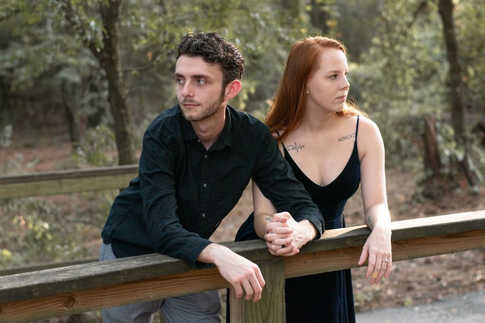 A couple in formal attire leans against a wooden railing in a park setting.