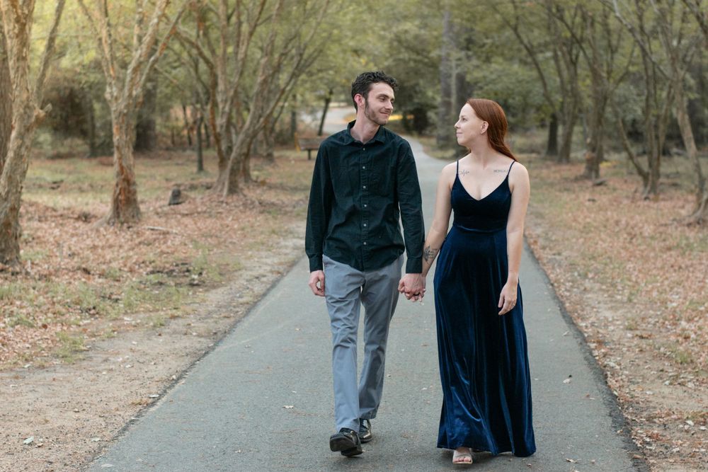 A couple walks hand in hand down a tree-lined path wearing formal attire.