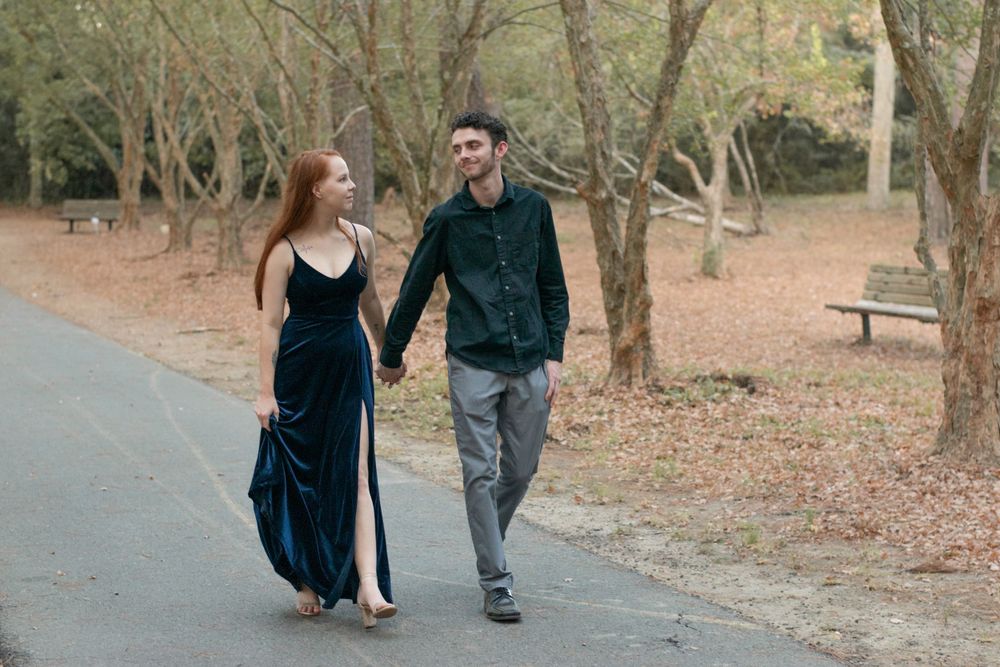 A couple strolls together on a paved pathway through a wooded park area.