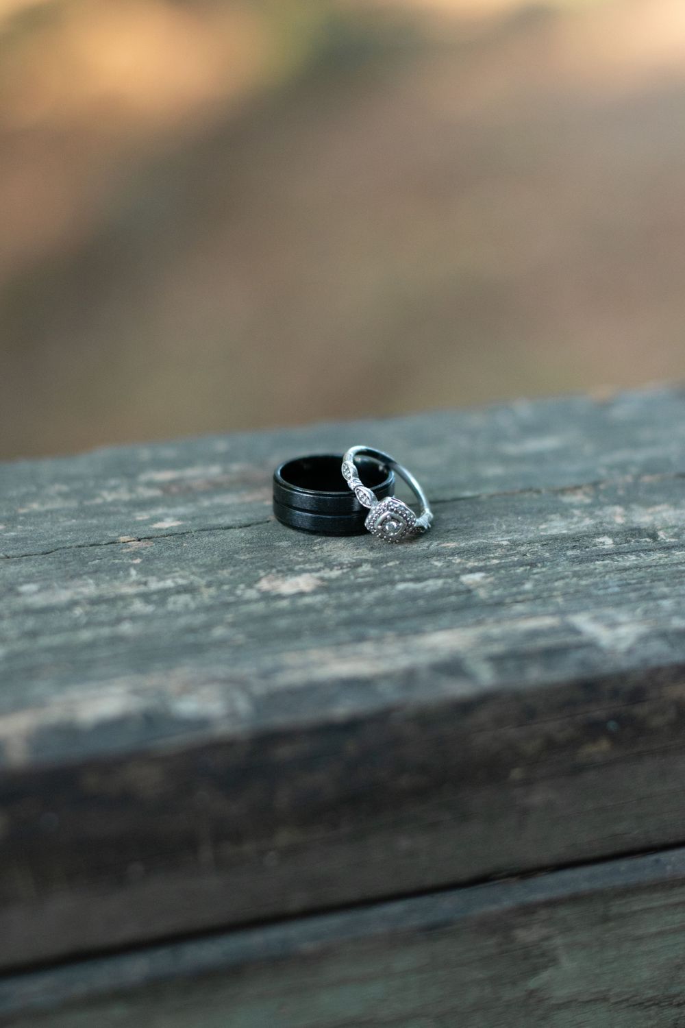 Two wedding rings, one diamond and one black band, sit on weathered gray wood in a close-up photo.