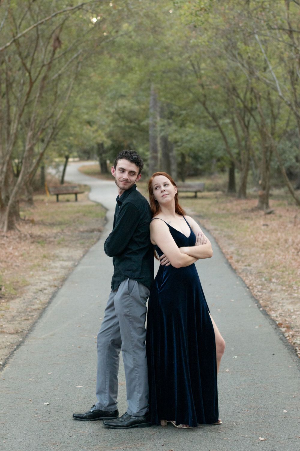 A couple poses back-to-back on a paved forest path wearing formal attire.