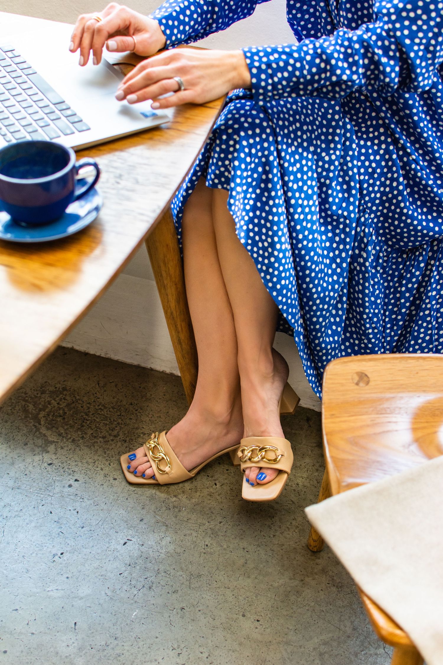 Someone wearing a polka dot blue dress and tan loafers works at a desk with a laptop and coffee cup.