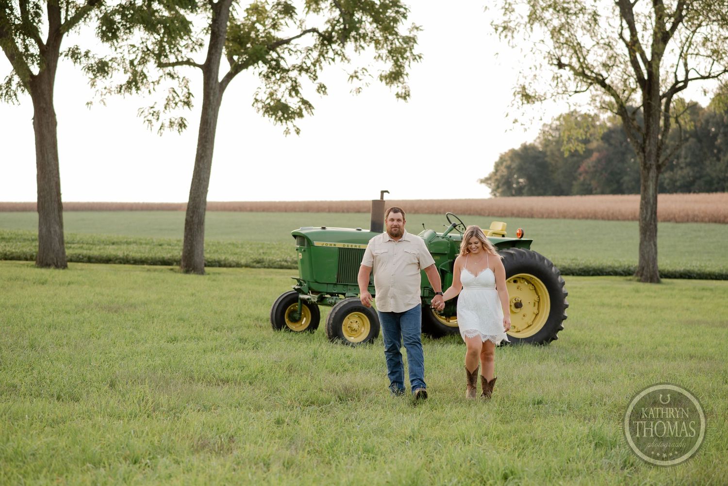 A couple poses together on a grassy field with a classic John Deere tractor in the background at sunset.