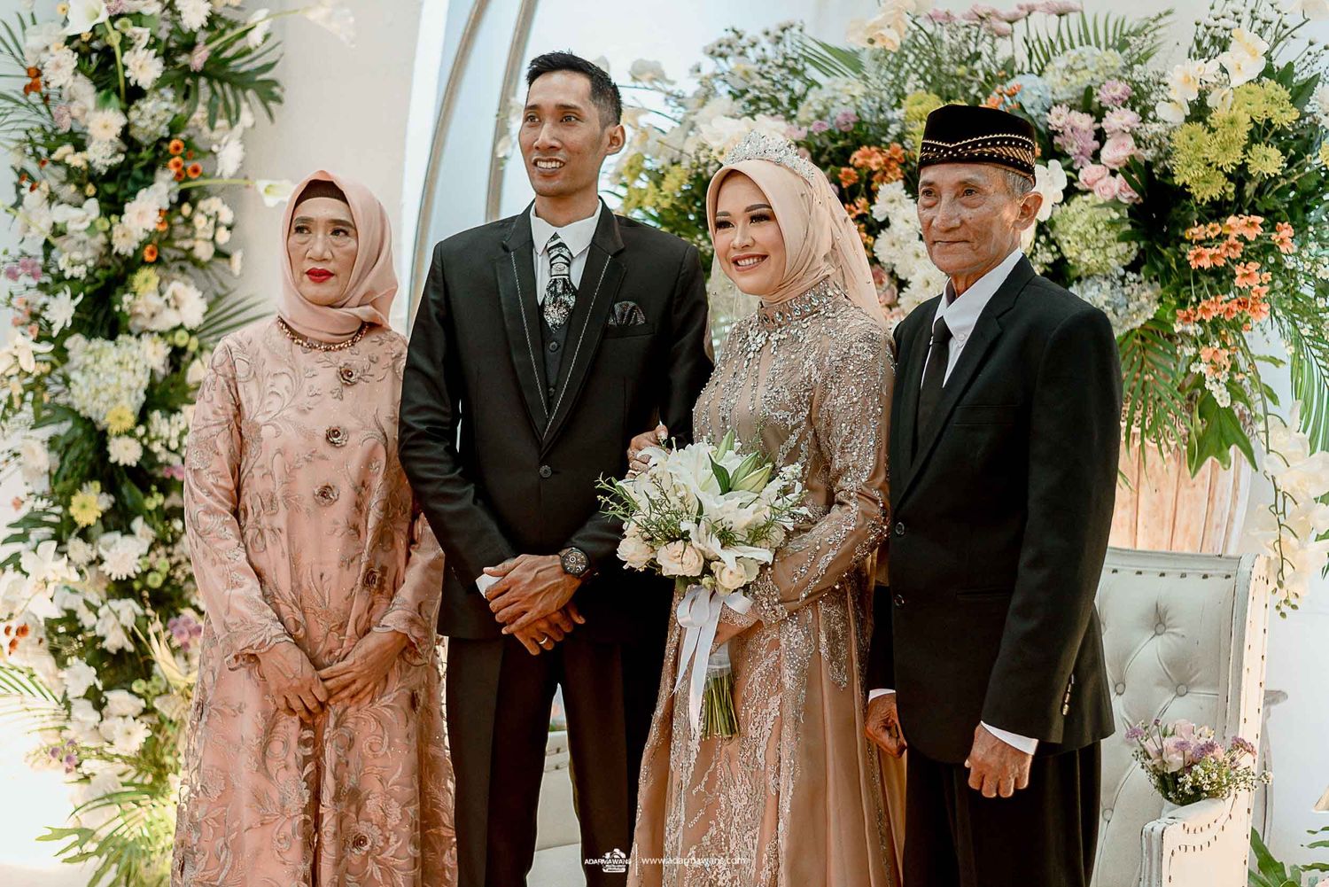 Wedding party stands together under floral arch with bride in sparkly dress holding white bouquet.