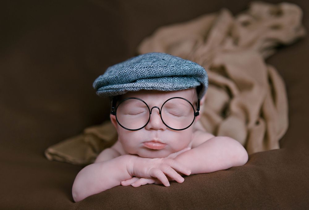 Vintage-styled newborn photo with baby wearing newsboy cap and glasses on brown backdrop.