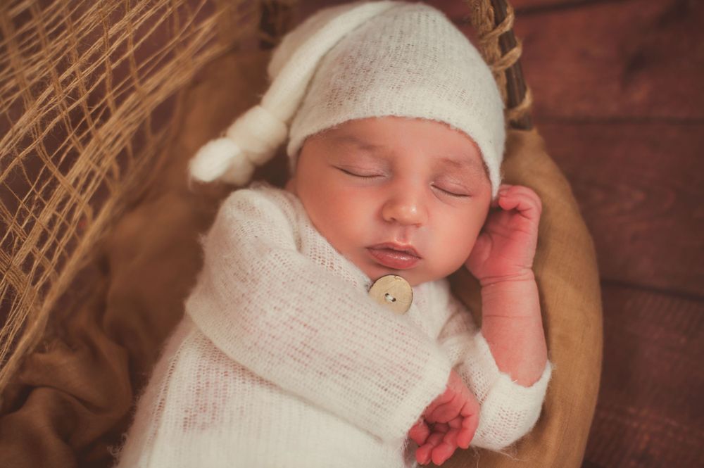 A sleeping newborn baby wearing a white knit outfit and hat in a wooden bowl.