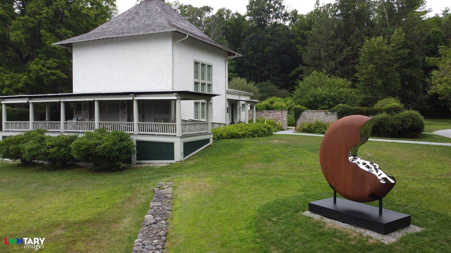 A curved metal sculpture sits in front of a white house with wraparound porch in a landscaped garden.